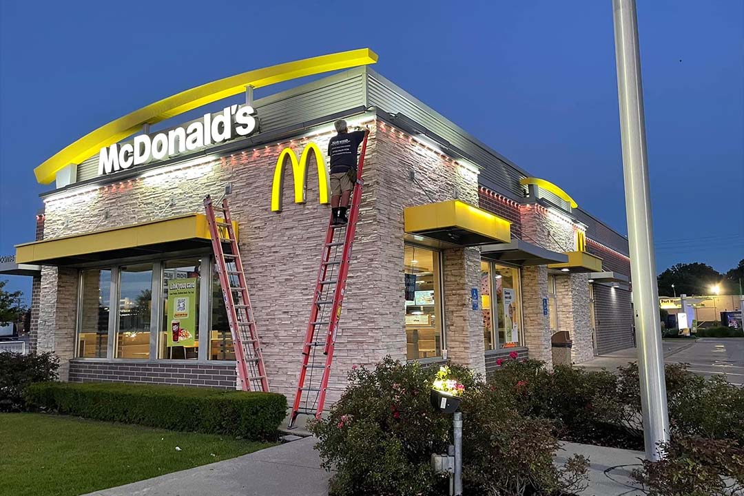 Attached image file 'n Werker op 'n leer verstel die ligte aan die buitekant van 'n McDonalds-restaurant teen skemer. Die gebou is verlig en die goue boë-logo is bo die ingang sigbaar.