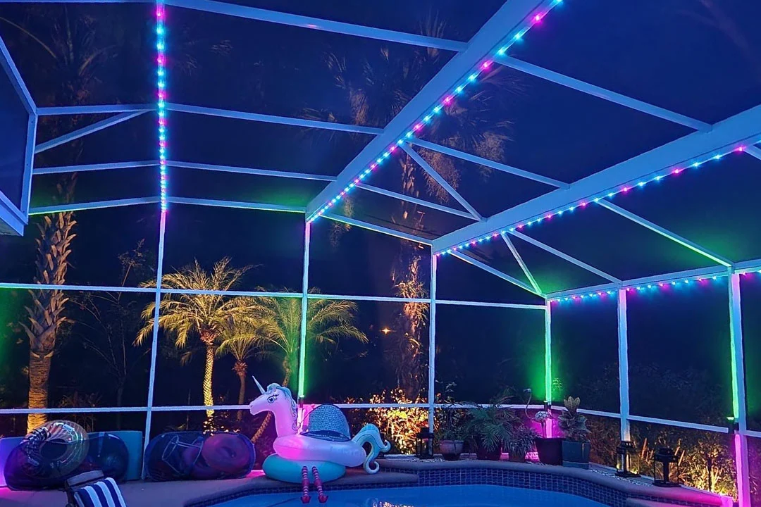 Attached image file 'n Afgeskermde patio word snags verlig met kleurvolle LED-ligte. Daar is palmbome buite, 'n eenhoring-swembadvlotter in die water, en verskeie patio-items en plante rondom die swembad.