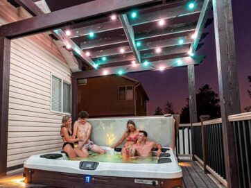 Vier mense in swemklere ontspan en gesels in 'n stomende warm bad op 'n dek onder 'n pergola met liggies, langs 'n huis in skemer.