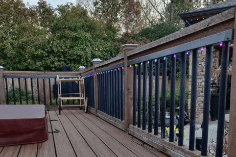 'n Houtdek met 'n reling versier met kleurvolle ligslingers kyk uit oor 'n tuin met digte groen bosse en bome. Patio-meubels en 'n opgevoude stoel is sigbaar op die dek.