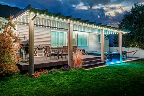 'n Agterplaas-patio met 'n pergola bedek met ligslingers, buitemeubels, 'n braai en 'n borrelbad, omring deur groen gras en landskappe, teen skemer.