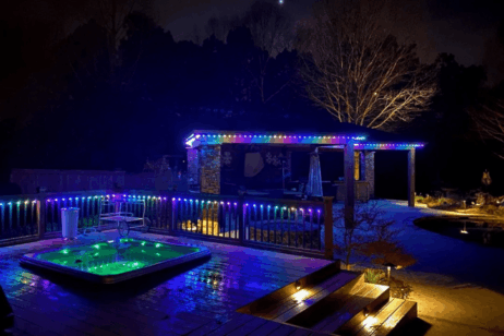 'n Agterplaas in die nag spog met 'n verligte borrelbad op 'n houtdek, wat met groen lig gloei. Kleurvolle LED-ligte omlyn die relings en 'n nabygeleë pergola, wat die buiteruimte teen 'n donker, boomryke agtergrond verlig.