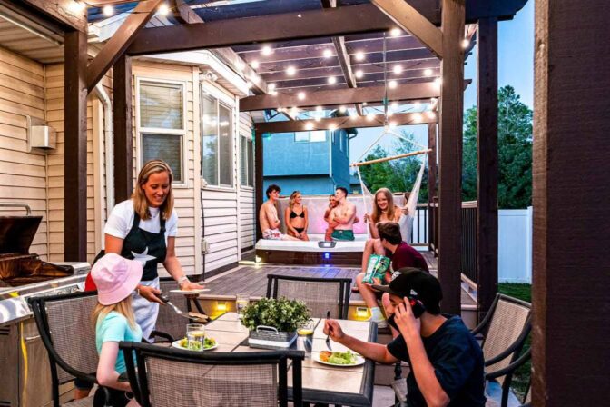 Attached image file Mense geniet 'n agterplaasbyeenkoms; sommige sit by 'n patiotafel en eet terwyl ander in 'n borrelbad onder liggies ontspan. Die toneel is lewendig en verwelkomend, met bome en 'n huis in die agtergrond.