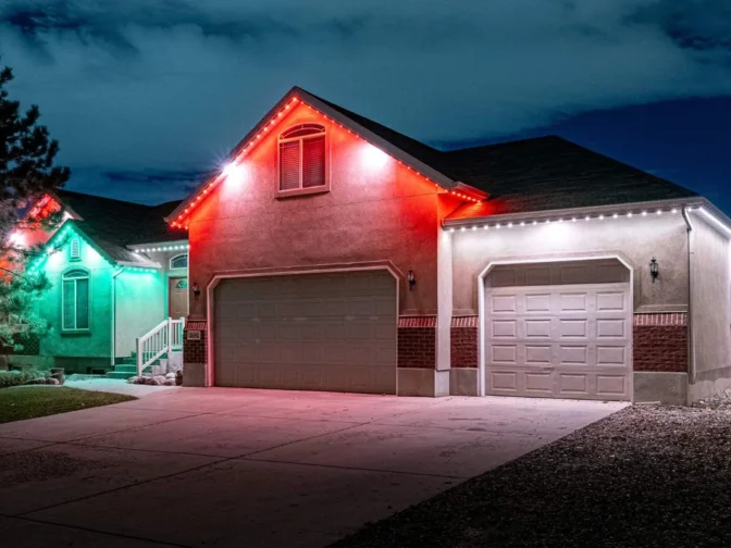 'n Enkelverdiepinghuis in die nag met sy dak omlyn in rooi en wit ligte, en 'n aangrensende huis met groen ligte, wat 'n feestelike vakansie-atmosfeer skep. Die lug is gedeeltelik bewolk.