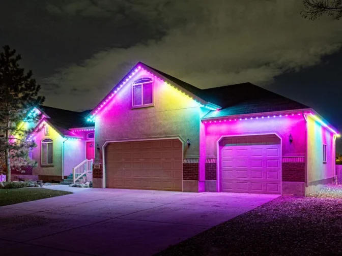 'n Huis in die nag met kleurvolle, helder LED-ligte wat die dak en motorhuis omlyn, wat in skakerings van pienk, pers, geel, groen en blou teen 'n bewolkte lug gloei.