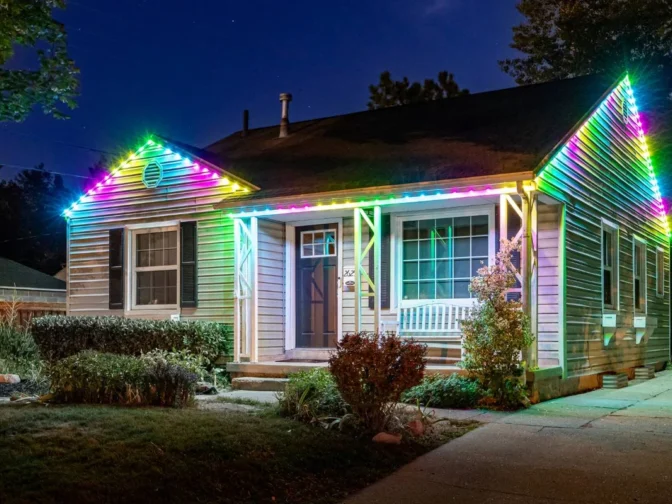 'n Enkelverdiepinghuis in die nag met sy daklyn versier met kleurvolle, gloeiende LED-ligte. Die huis het ligte gevelbekleding, 'n voorstoep en 'n klein tuin. Die vensters word van binne verlig.