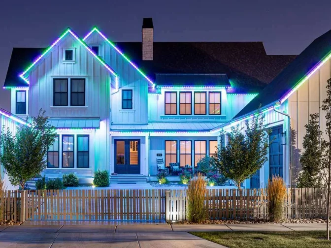 'n Moderne tweeverdiepinghuis in die nag, versier met blou, groen en pers LED-ligte langs die daklyn. Die huis het groot vensters, 'n voorstoep en 'n wit heining met 'n paar bome en struike in die erf.