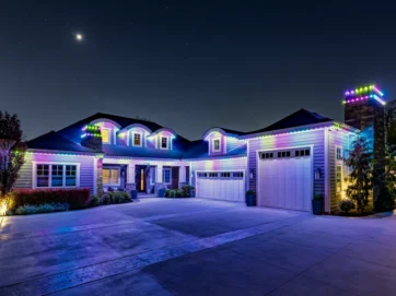 'n Groot huis is snags versier met kleurvolle LED-ligte wat die dak, motorhuis en skoorsteen omlyn. Die oprit is leeg, en die maan en sterre is sigbaar in die donker lug bo.
