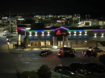 'n Chick-fil-A-restaurant in die nag, helder verlig met kleurvolle ligslingers langs die dak, omring deur geparkeerde motors en 'n besige deurry. Stadsligte en ander besighede is in die agtergrond sigbaar.