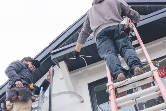 Attached image file Twee werkers op lere installeer vakansieligte langs die daklyn van 'n huis; een hou snaarligte vas terwyl die ander gereedskap gebruik en 'n gereedskapsgordel dra.