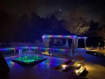 'n Agterplaas in die nag beskik oor 'n houtdek met 'n borrelbad verlig deur kleurvolle LED-ligte, 'n pergola met bypassende ligte en 'n klein verligte paadjie langs 'n swembad, alles omring deur bome onder 'n donker lug.