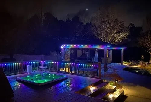 Attached image file 'n Agterplaas in die nag beskik oor 'n houtdek met 'n borrelbad verlig deur kleurvolle LED-ligte, 'n pergola met bypassende ligte en 'n klein verligte paadjie langs 'n swembad, alles omring deur bome onder 'n donker lug.