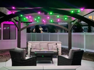 Buitepatio met 'n pergola bo-oor, versier met pienk en groen ligslingers. Die area beskik oor 'n gestoffeerde bank, twee rietstoele en 'n tafel, omring deur 'n wit privaatheidsheining teen skemer.