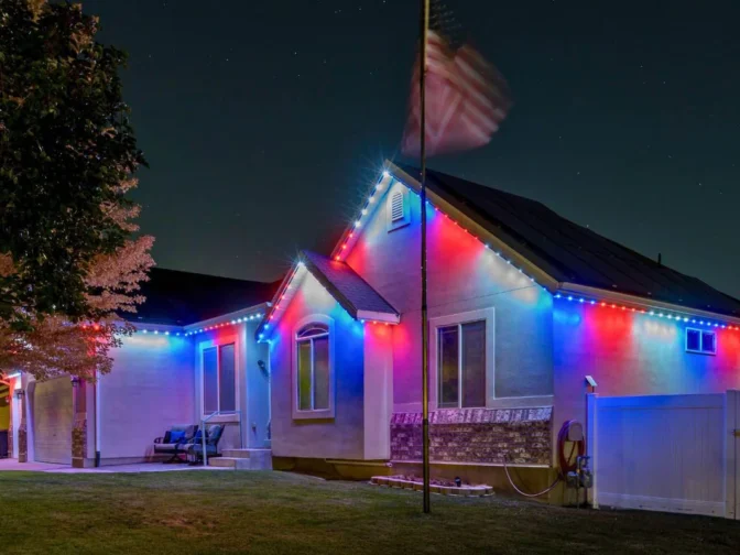 'n Huis is snags versier met rooi, wit en blou ligte langs die daklyn. 'n Amerikaanse vlag wapper op 'n paal in die voortuin, en 'n boom en wit heining is sigbaar.
