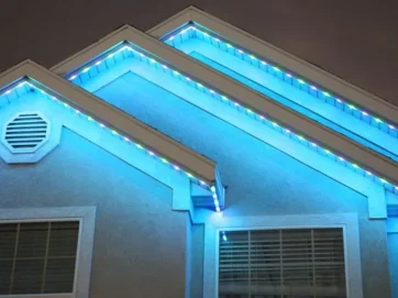 Die boonste gedeelte van 'n huis in die nag vertoon gloeiende blou en wit ligte wat die dakrande omlyn en die driehoekige vorms bo twee vensters uitlig.