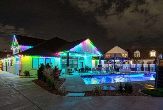 Attached image file 'n Moderne huis word snags verlig met kleurvolle LED-ligte langs die daklyn. Die agterplaas beskik oor 'n swembad, leunstoele en 'n pergola, wat 'n lewendige en uitnodigende atmosfeer skep.