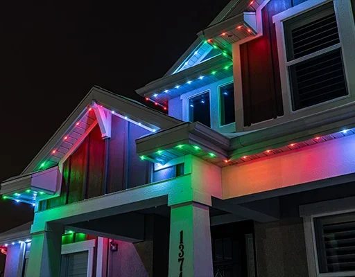 Attached image file 'n Tweeverdiepinghuis in die nag versier met veelkleurige LED-ligte langs die daklyne, wat die buitekant met rooi, groen en blou skakerings verlig. Huisnommer 137 is sigbaar op 'n pilaar.