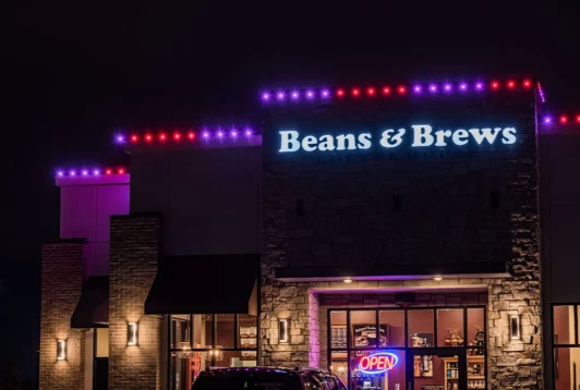 Attached image file 'n Beans & Brews-koffiewinkel in die nag, met pers en rooi ligte wat die dak omlyn en 'n helder neon OOP-teken wat in die voorvenster gloei.