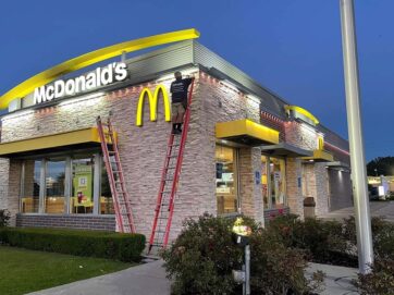 'n Werker op 'n leer verstel die ligte aan die buitekant van 'n McDonalds-restaurant teen skemer. Die gebou is verlig en die goue boë-logo is bo die ingang sigbaar.