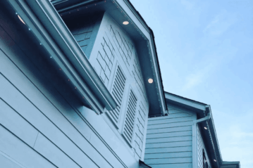 Naby-aansig van die buitekant van 'n moderne blou huis met horisontale gevelbekleding, 'n dakvenster met toe luike, ingeboude ligte en 'n helderblou lug in die agtergrond.