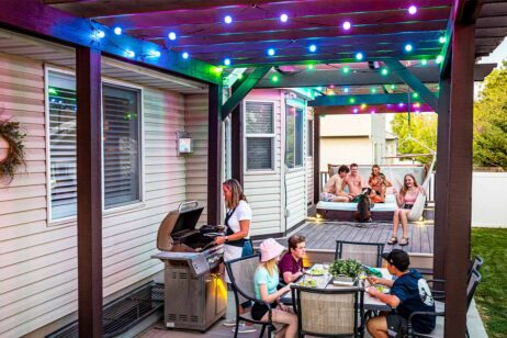 Mense geniet 'n agterplaasbyeenkoms met sonsondergang. 'n Vrou braai kos terwyl kinders by 'n tafel eet. Meer mense ontspan op 'n verhewe dek met kleurvolle ligslingers en 'n hond, wat 'n lewendige, feestelike atmosfeer skep.