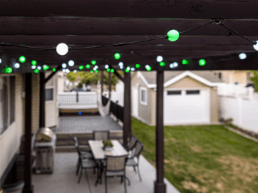 Ligslingers met groen en wit gloeilampe hang oor 'n oordekte patio met 'n eetkamertafel en stoele. In die agtergrond is daar 'n klein grasperk, 'n losstaande motorhuis en 'n deel van 'n dek met 'n wit reling.