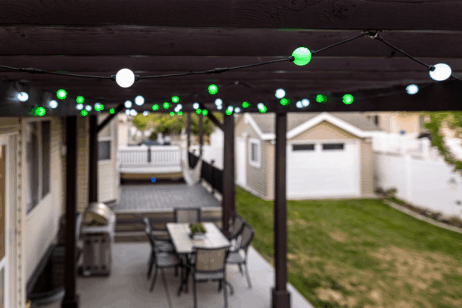 Ligslingers met groen en wit gloeilampe hang oor 'n oordekte patio met 'n eetkamertafel en stoele. In die agtergrond is daar 'n klein grasperk, 'n losstaande motorhuis en 'n deel van 'n dek met 'n wit reling.