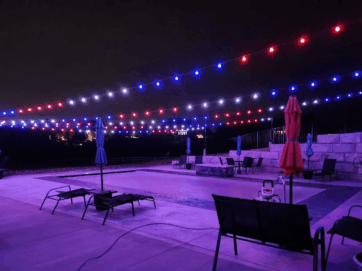 'n Patio langs die swembad word snags verlig deur stringe rooi, wit en blou ligte. Ligstoele en sambrele is rondom die swembad gerangskik, met klipmure in die agtergrond. Die toneel het 'n feestelike atmosfeer.