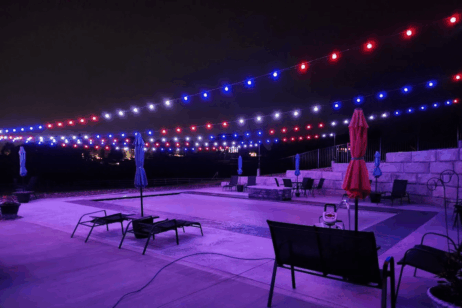 'n Patio langs die swembad word snags verlig deur stringe rooi, wit en blou ligte. Ligstoele en sambrele is rondom die swembad gerangskik, met klipmure in die agtergrond. Die toneel het 'n feestelike atmosfeer.