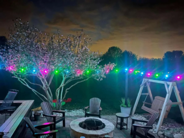 'n Agterplaas-patio in die nag met Adirondack-stoele rondom 'n vuurput, 'n houtswaai, blombome en kleurvolle ligslingers wat in die agtergrond onder 'n bewolkte lug gloei.