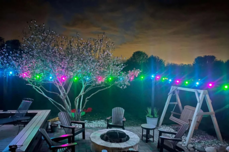 'n Agterplaas-patio in die nag met Adirondack-stoele rondom 'n vuurput, 'n houtswaai, blombome en kleurvolle ligslingers wat in die agtergrond onder 'n bewolkte lug gloei.