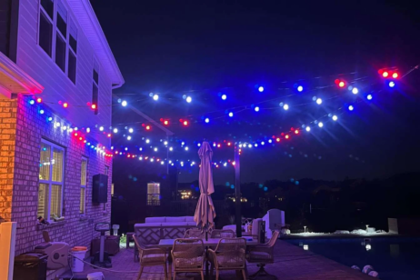 'n Patio in die nag met rooi, wit en blou ligsnoere wat bo buitemeubels hang, naby 'n swembad langs 'n baksteenhuis. Die ligte verlig die area en skep 'n feestelike atmosfeer.