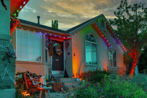 'n Huis versier vir Halloween teen skemer, met kleurvolle liggies, spinnekoppe en vals spinnekoppe op die stoep. Twee stoele en Halloween-bykomstighede is naby die voorstoep gerangskik, omring deur plante en blomme.