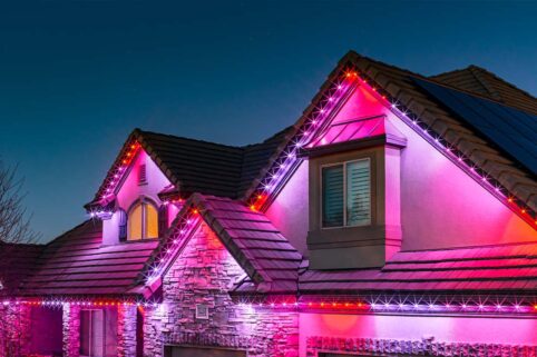'n Moderne huis teen skemer, daklyne omlyn met helderpienk, rooi en wit LED-ligte, wat die klip- en pleisterwerk-buitekant teen 'n helder aandhemel verlig.