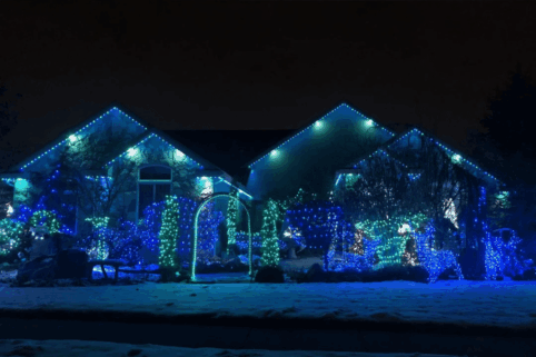 'n Huis en erf versier met blou en groen vakansieliggies in die nag, met bome en bosse omlyn in ligte. Sneeu bedek die grond en skep 'n feestelike wintertoneel.