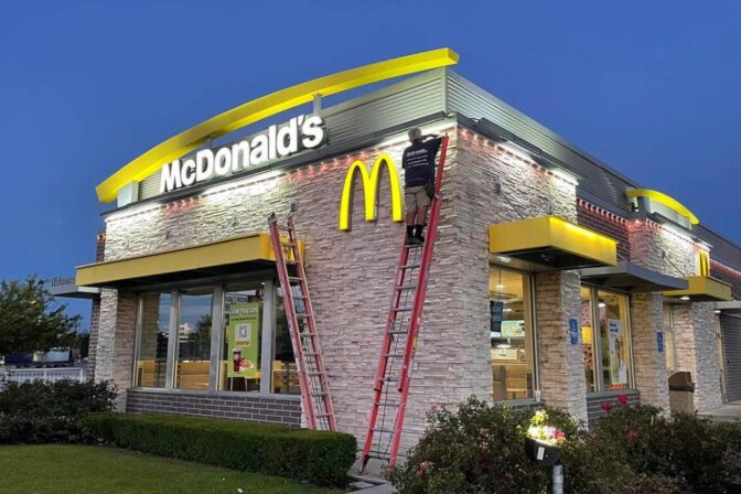 Attached image file 'n Werker staan op 'n leer en verstel rooi vakansieligte bo die ingang van 'n McDonalds-restaurant teen skemer. Nog 'n leer leun teen die gebou, en buiteligte en -tekens is duidelik sigbaar.