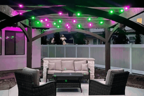 Buitepatio met 'n beige bank, twee donker rietstoele, 'n koffietafel en ligsnoere wat pienk en groen gloei onder 'n houtpergola, teen 'n wit heining en aangrensende huise.