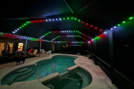 'n Agterplaas swembad in die nag onder 'n oordekte patio, verlig deur stringe rooi, groen en wit LED-ligte. Mense sit naby die swembad, en 'n hond staan naby. Die water weerkaats die kleurvolle ligte hierbo.