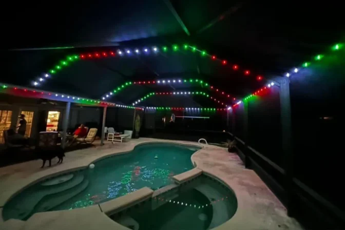 Attached image file 'n Agterplaas swembad in die nag onder 'n oordekte patio, verlig deur stringe rooi, groen en wit LED-ligte. Mense sit naby die swembad, en 'n hond staan naby. Die water weerkaats die kleurvolle ligte hierbo.