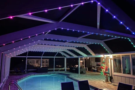 'n Oordekte buitepatio met 'n swembad is versier met liggies in pers, blou en groen. Patio-meubels en 'n verligte Kersboom is snags naby die huis sigbaar.