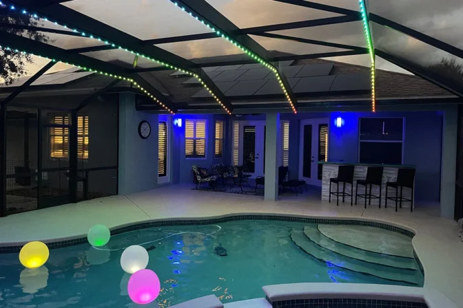 Attached image file 'n Swembadarea in die agterplaas teen skemer met gloeiende, kleurvolle drywende balle in die water en veelkleurige LED-strookligte op die patio-dak. Patio-meubels en kroegstoele is in die agtergrond sigbaar.