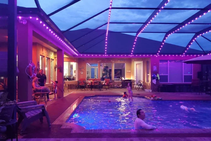 Attached image file Mense geniet 'n aandse swembadpartytjie onder 'n beskutte patio met pers liggies. Sommige swem, terwyl ander by die swembad sit en gesels. Die huis en patio is warm verlig, wat 'n feestelike atmosfeer skep.