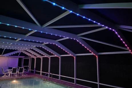 'n Oordekte patio met 'n swembad en patiomeubels, verlig deur LED-strookligte in blou, pers en rooi, wat die balke van die dak snags omlyn.