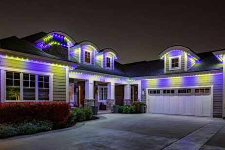 'n Moderne huis in die nag word omlyn met pers, blou en wit LED-ligte langs die dak en dakvensters, wat 'n feestelike en lewendige voorkoms skep. Die oprit en voortuin is sigbaar in die voorgrond.
