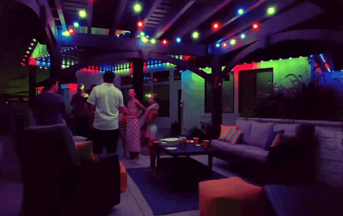 Attached image file Mense kom bymekaar en gesels onder 'n pergola versier met kleurvolle ligslingers in die nag. Patio-meubels en tafels met drankies en kos is sigbaar, wat 'n lewendige, feestelike agterplaasatmosfeer skep.