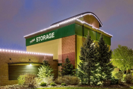 'n Moderne bergingsfasiliteit met 'n groen en wit TH!NK STORAGE-teken, 'n baksteen- en bruin buitekant, verligte versieringsligte en aangelegde bome en bosse voor, gefotografeer teen skemer.